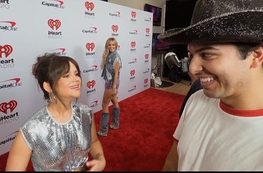 Ricky Garcia on the iHeartCountry Festival red carpet, mid-conversation with an artist, a microphone in frame.